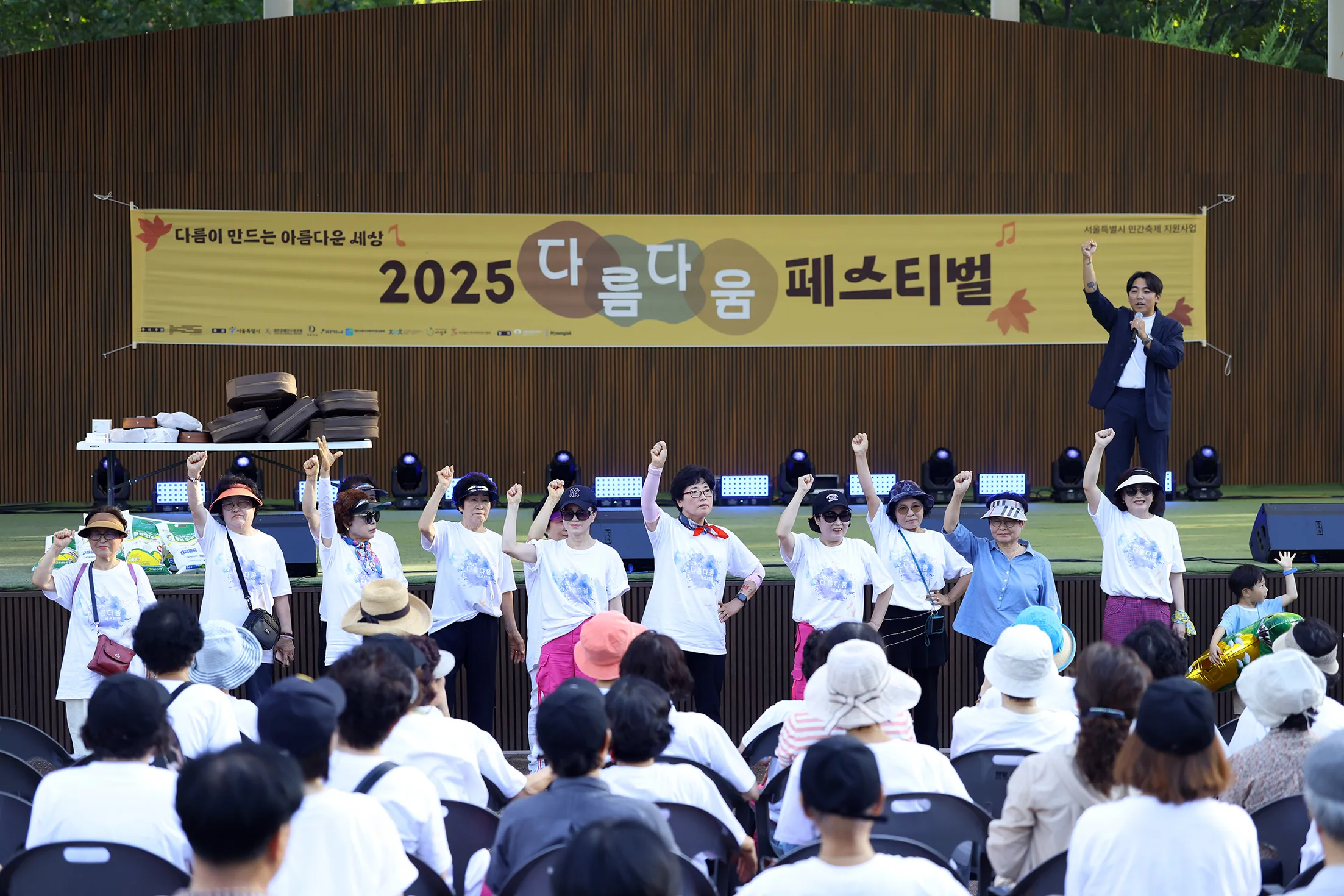 '2025 다름다움 페스티벌 — 다름이 만드는 아름다운 세상' 현수막이 걸린 무대 앞에 흰 티셔츠를 맞춰 입은 참가자 열두어 명이 한 줄로 서서 한쪽 주먹을 치켜들고 있다. 무대 위 진행자도 같은 동작을 하고, 뒤편 객석은 관객으로 가득 차 있다.